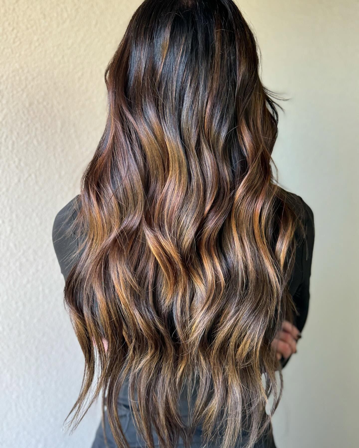 Long, wavy, dark brown hair with warm caramel-toned balayage highlights, viewed from behind.